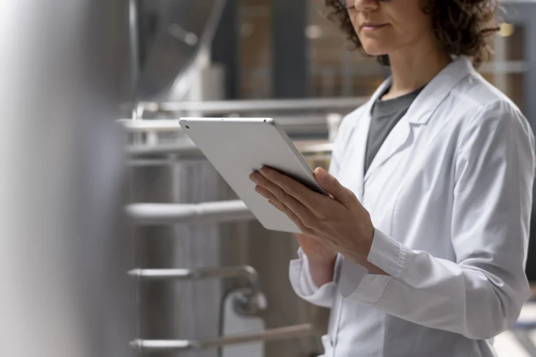 Femme de profil, en blouse blanche, avec une tablette tactile à la main en train de contrôler un laboratoire